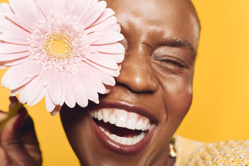 Happy African woman with dark skin laughing and holding pink flower near her eye on yellow background. Concept of joy, beauty, skincare, and natural happiness in studio lighting.