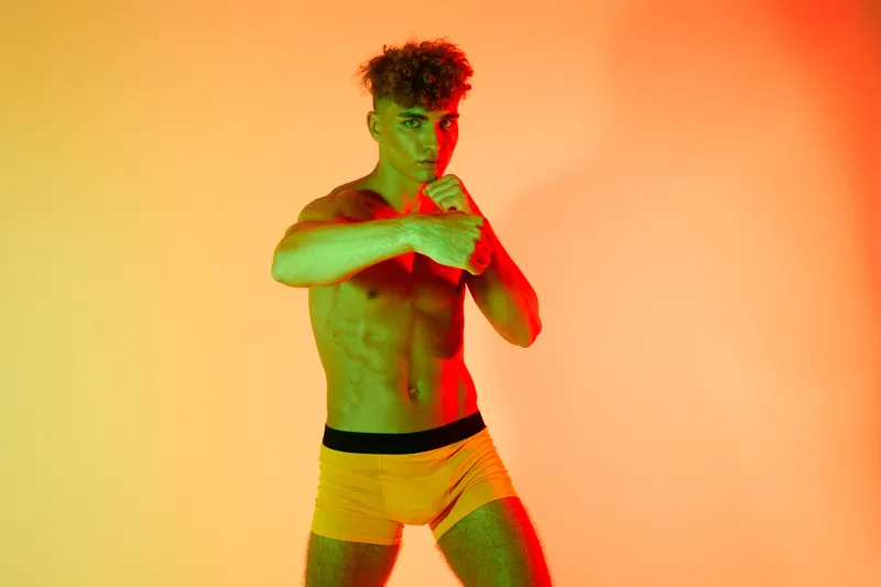 Young man with muscular body posing in fighting stance wearing shorts with red and green gradient light on orange background for fitness sport motivation and boxing training.
