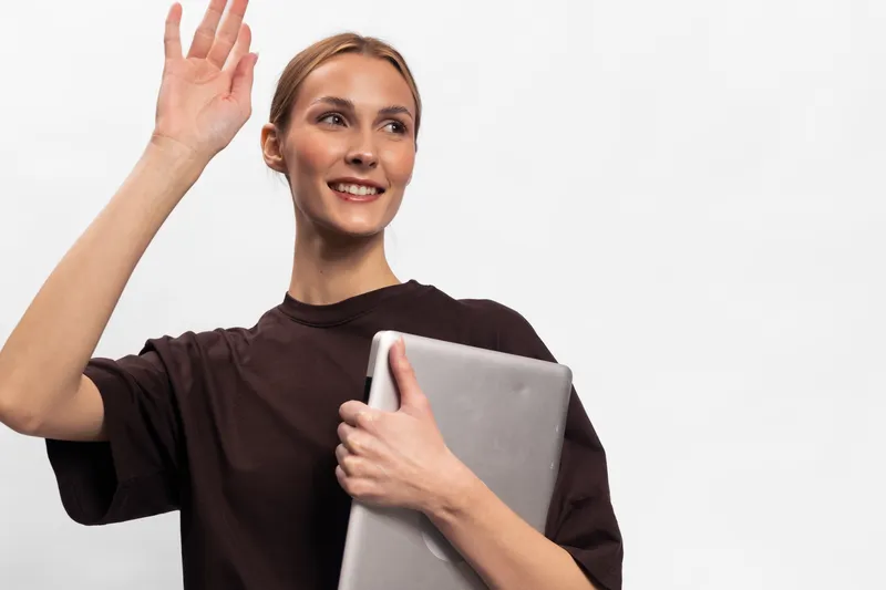 Smiling young woman holding laptop waves hello with raised hand, wearing casual dark shirt. Isolated on white background. Concept for greeting, technology, communication, and remote work.