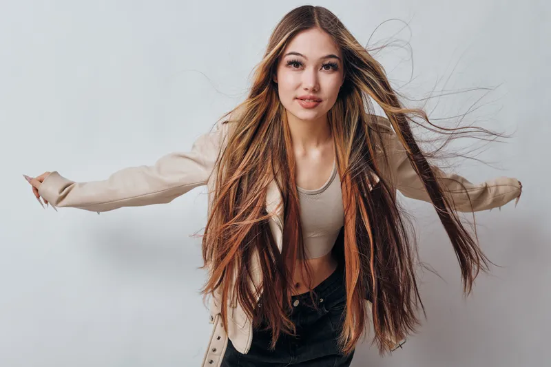 Young woman with long brown hair wearing light jacket and top posing with arms outstretched against white background for fashion or lifestyle.