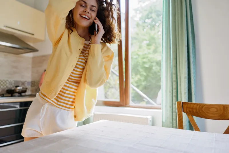 A young woman in a yellow cardigan talks on her phone in a bright kitchen, smiling beside a wooden table.