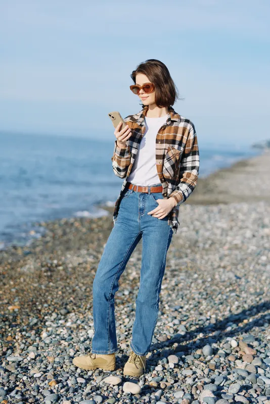 Casual woman on a pebble beach checks her phone, wearing a plaid shirt, jeans and boots, enjoying a sunny seaside moment with a light ocean breeze