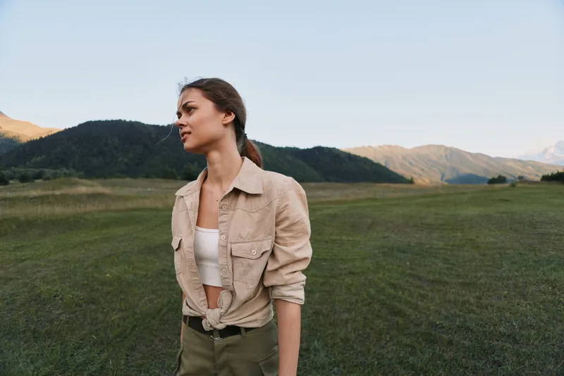 Young woman with brunette hair stands thoughtfully in a vast green field with mountains in the background during a clear day, wearing a beige jacket and white top.