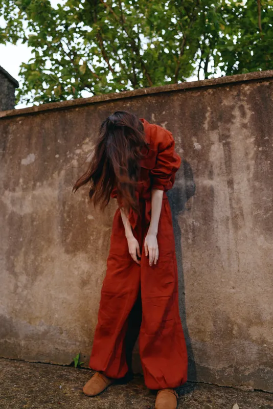 A young woman in a red jumpsuit bends beside a weathered wall, creating a moody street scene that captures candid fashion, urban resilience, and a bold, confident vibe