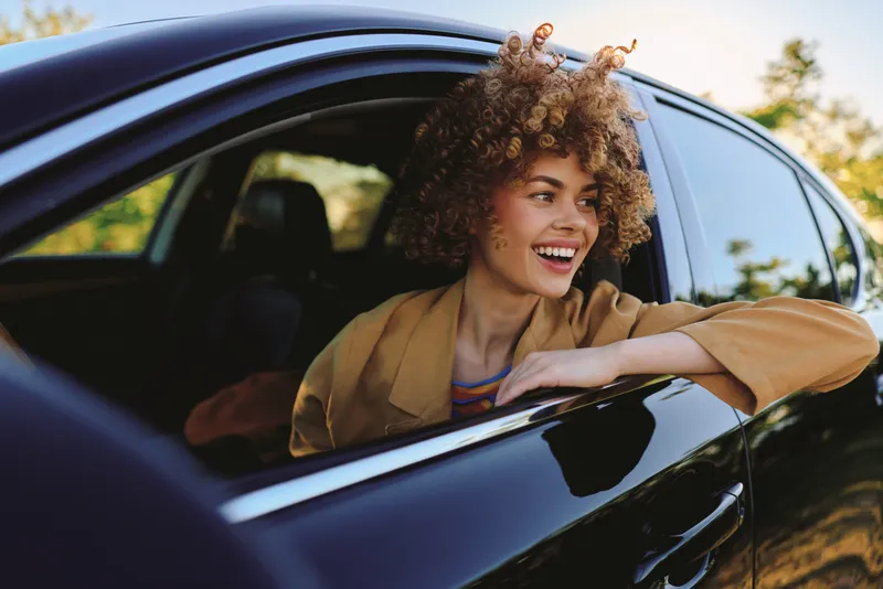 Smiling woman leaning from a sleek car window, curly hair catching sunlight. Outdoor scene suggesting carefree lifestyle, travel, and self expression. Retro film color tone enhances a warm, nostalgic