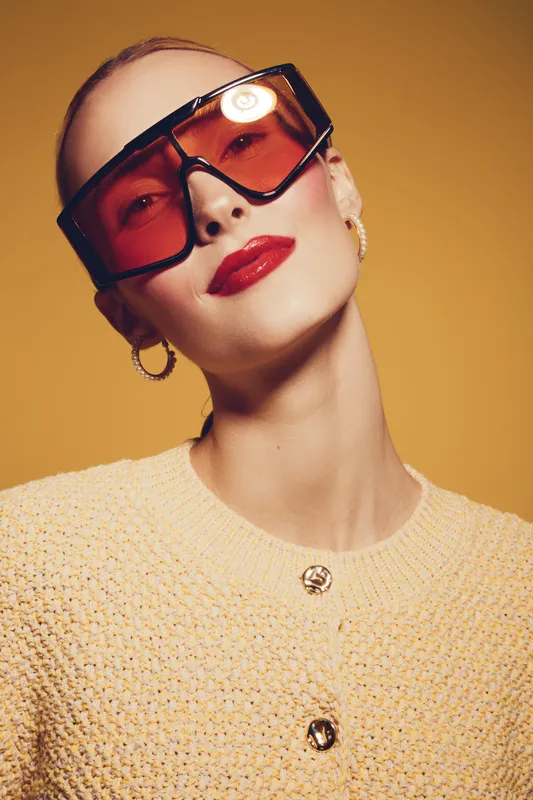 Stylish woman with slicked back hair wears oversized red sunglasses and gold hoop earrings. She poses confidently in a textured beige sweater against a warm yellow background.