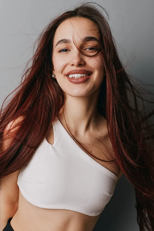 Young woman with long hair smiling and looking at camera wearing white crop top against gray background. Casual portrait with natural makeup and joyful expression indoors.
