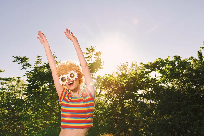 Playful woman wearing a striped top and flower sunglasses jumps joyfully in a sunny park, capturing a lively lifestyle moment with retro film color and carefree mood outdoors under bright light.