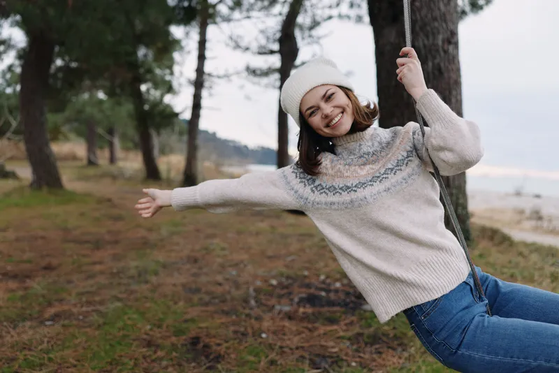Woman on a swing in a snowy woodland by a lake, wearing a cozy knit sweater and beanie, smiling as she enjoys outdoor winter leisure among pine trees