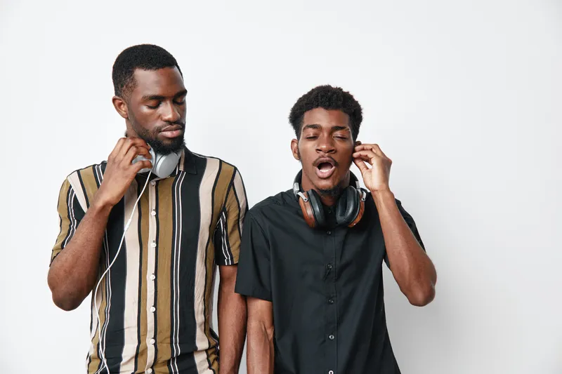 Two young black men with dark skin listening to music with headphones, one wearing striped shirt and the other in black shirt, standing against white background indoors.
