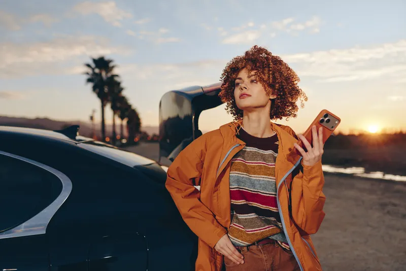 Curly haired woman standing by car at sunset, holding smartphone, casual outdoors lifestyle, warm golden sky, palm trees in distance, evening light, relaxed travel portrait today
