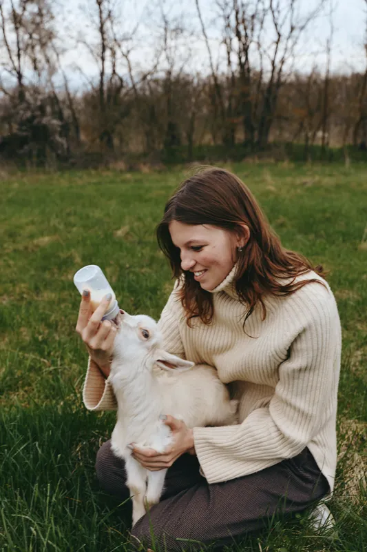 Lifestyle scene of a woman outdoors feeding a goat kid with a bottle, smiling softly, cozy sweater, natural light, lush grass, peaceful countryside, warm connection, candid moment