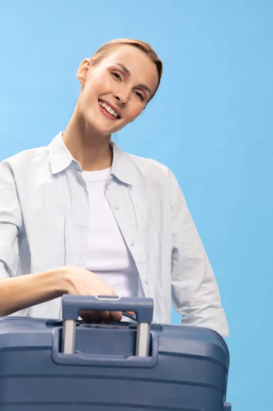 Smiling woman in casual white shirt holding blue suitcase on blue background. Travel concept with happy female traveler ready for trip, vacation, or business journey. Positive mood, studio lighting.