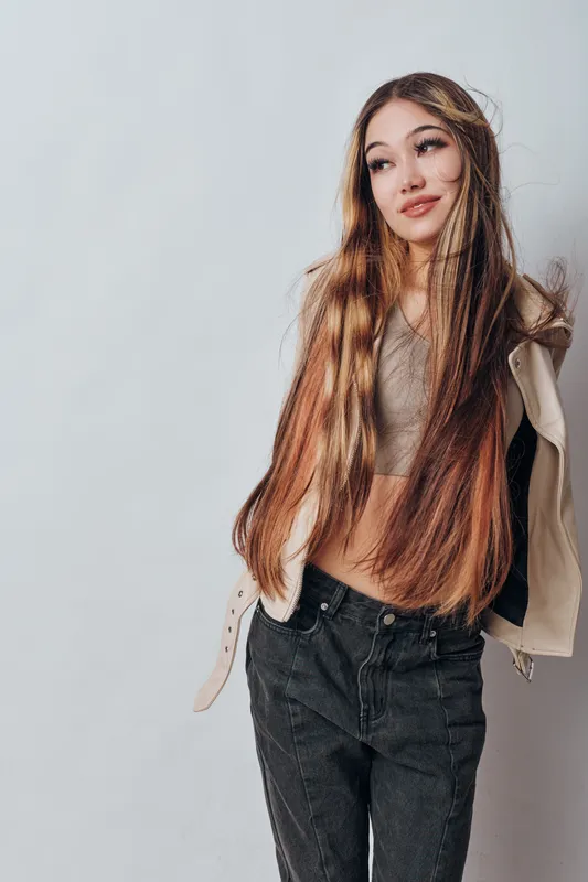 Young woman with long multicolored hair posing in casual clothing with jacket and jeans looking away from camera on white background indoors.