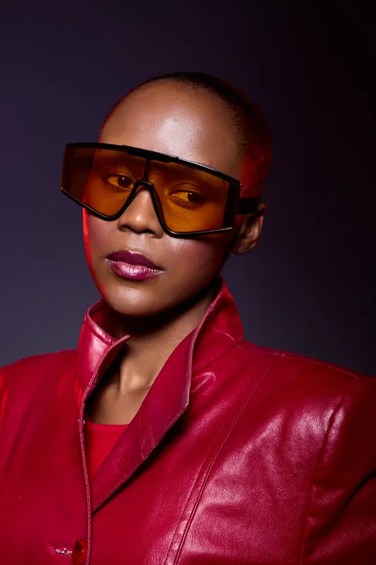 Stylish African American woman with dark skin wearing large orange sunglasses and a red leather jacket poses against dark background. Concept of fashion, confidence, and modern style.