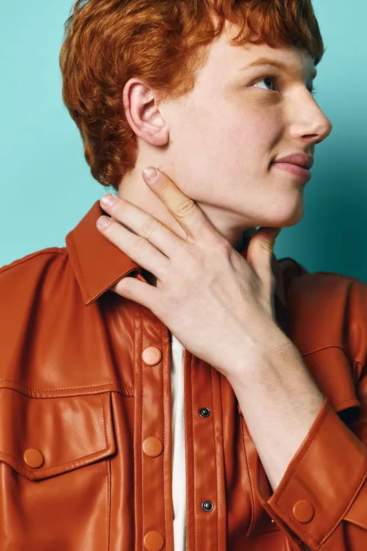 Young redhead man in brown leather jacket touching his neck with hand, looking sideways against blue background in studio portrait, casual fashion expression.