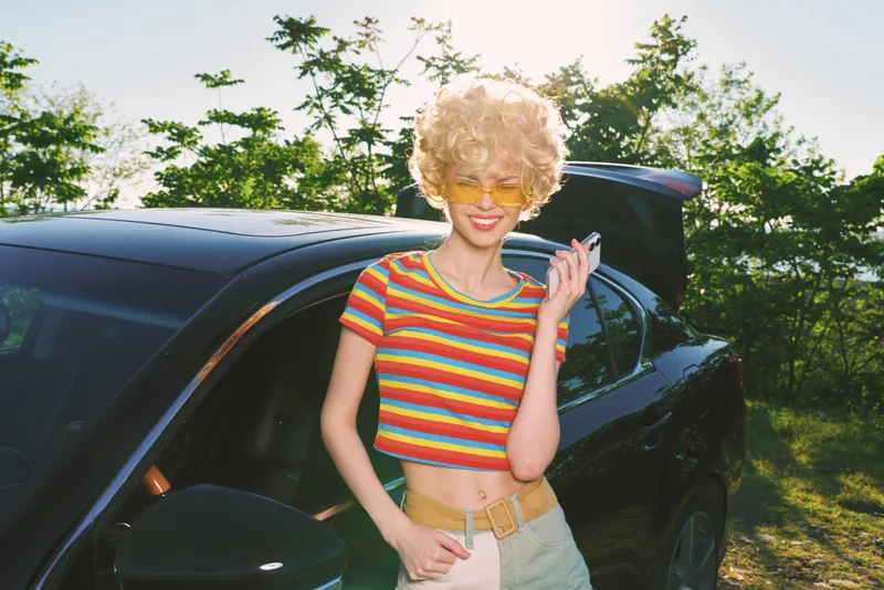 Cheerful woman with curly blonde hair wearing a striped crop top stands beside a car in bright outdoor light, conveying casual lifestyle, energetic wellness, and retro film color.