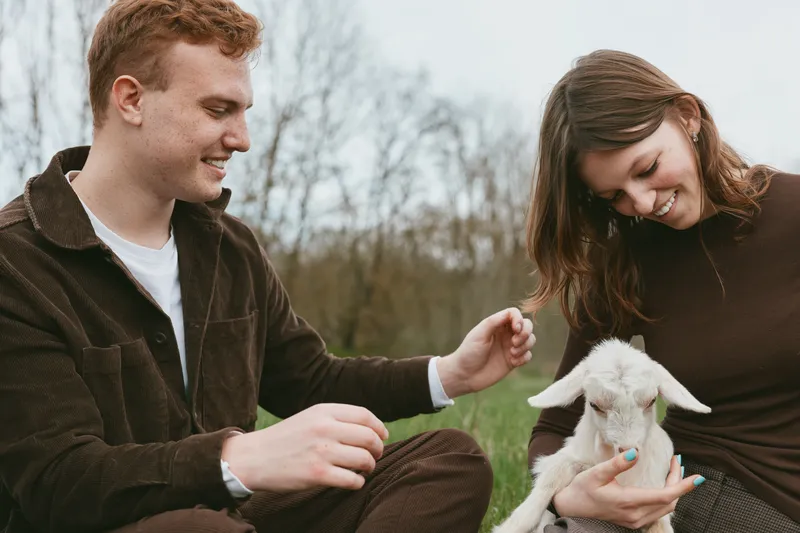 Cheerful couple outdoors sharing a light moment with a playful goat kid, relaxed posture, natural colors, grassy field, gentle breeze, warm connection, lifestyle scene.