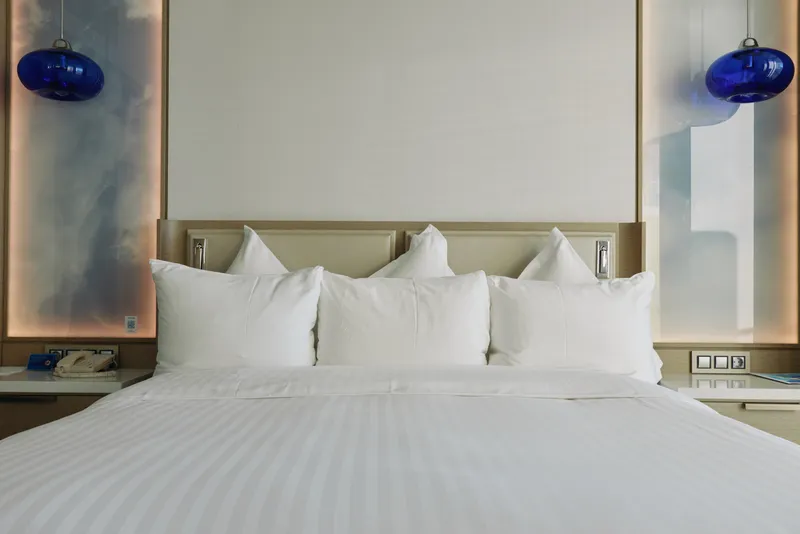 Bed, pillows and pristine white linens dominate a sleek hotel room with soft lighting, plush comfort and modern headboard, inviting restful sleep and daytime lounging in a calm, luxurious space