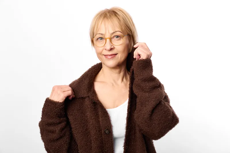 Smiling older woman with glasses, studio portrait on white background, brown fuzzy jacket over white top, hands raised in confident pose, soft neutral light, clean backdrop