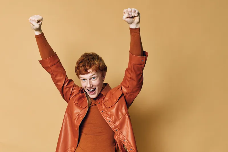 Young man with red hair wearing brown leather jacket and sweater raises fists up celebrating success on beige background. Portrait, excited mood, casual style and energetic emotion.