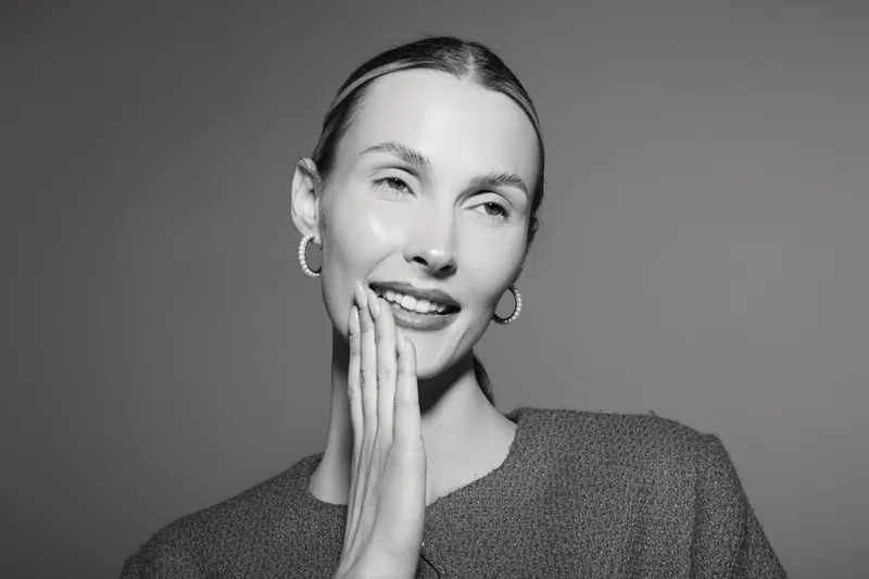 Smiling Caucasian woman with light skin touches her face gently, wearing hoop earrings and a textured top. Studio portrait with soft lighting and minimalistic background.