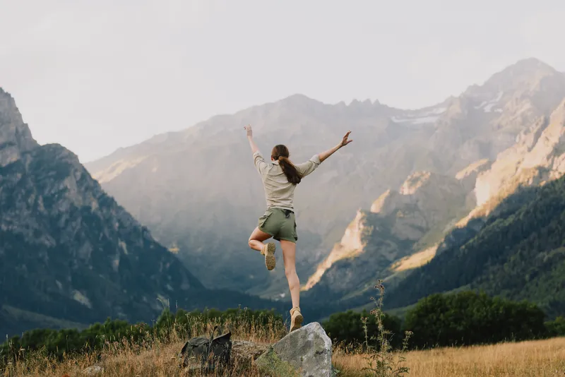 Young woman leaps on a rock in an alpine valley with towering mountains behind, radiating freedom and energy in an outdoor scene that feels spontaneous and lively