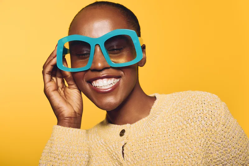 Smiling African woman with dark skin wearing large blue sunglasses and yellow sweater poses against bright yellow background. Concept of fashion, summer style, and joyful mood.