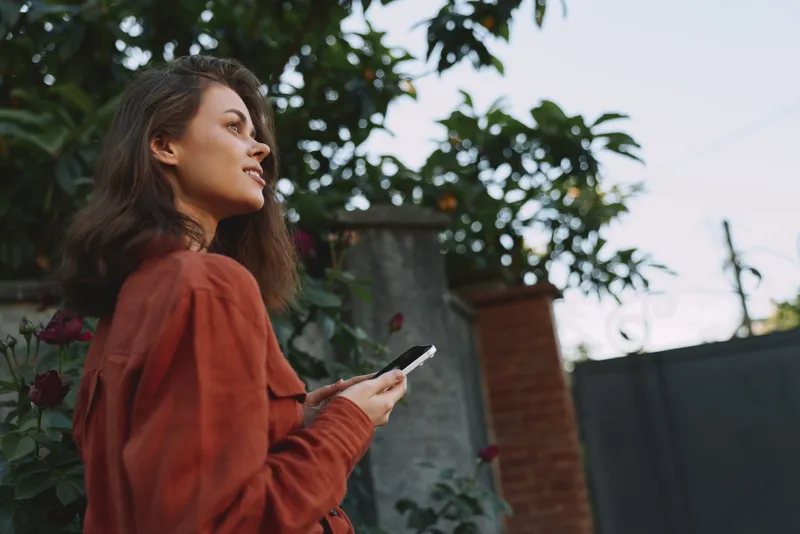 Woman in a red sweater stands outdoors, looking to the side, holding a smartphone. Natural light highlights a relaxed lifestyle, casual fashion, and a candid moment
