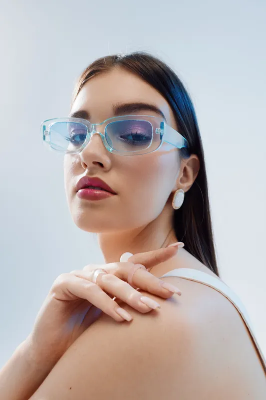 Young woman with long dark hair wearing transparent glasses and white earrings poses confidently against a light background. Concept of beauty, fashion, and modern style.