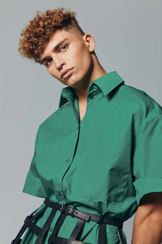 Young man with curly hair wearing green shirt and black belt posing against gray background in studio for fashion portrait and style concept.
