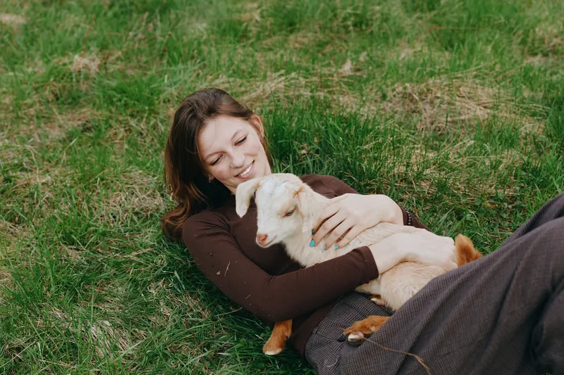 Cheerful woman outdoors, lying on grass with a playful goat kid, warm sunlight, relaxed pose, soft breeze, natural colors, meadow backdrop, the moment feels carefree and cozy