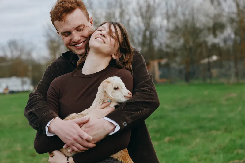A joyful couple embraces a small goat in an open field, capturing warmth, affection, and a relaxed rural lifestyle under soft natural light, creating a calm, loving mood.