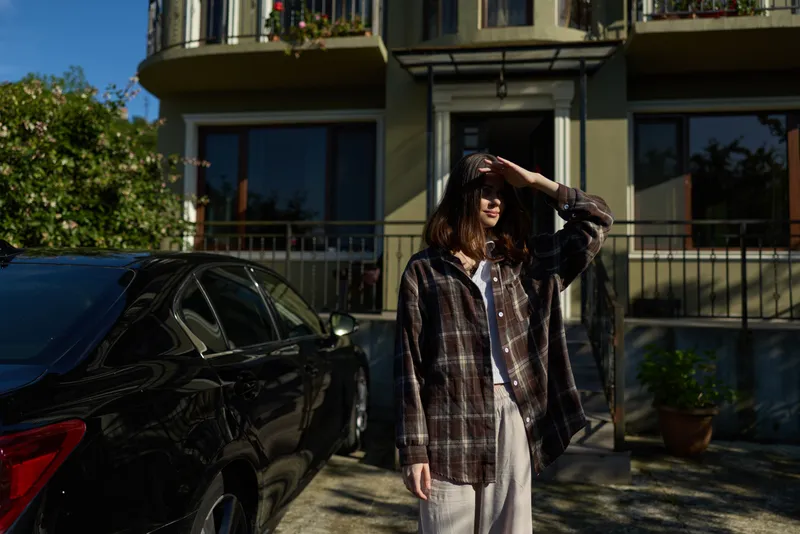 Casual woman stands beside a car on a sunny outdoor street, wearing a plaid shirt and relaxed jeans, embodying lifestyle and casual fashion in a candid urban moment