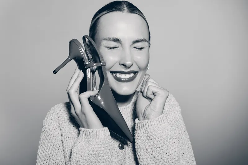 Happy caucasian woman with fair skin in knitted sweater holds high heel shoe near face smiling with closed eyes in studio. Concept of fashion, joy, and lifestyle for women.