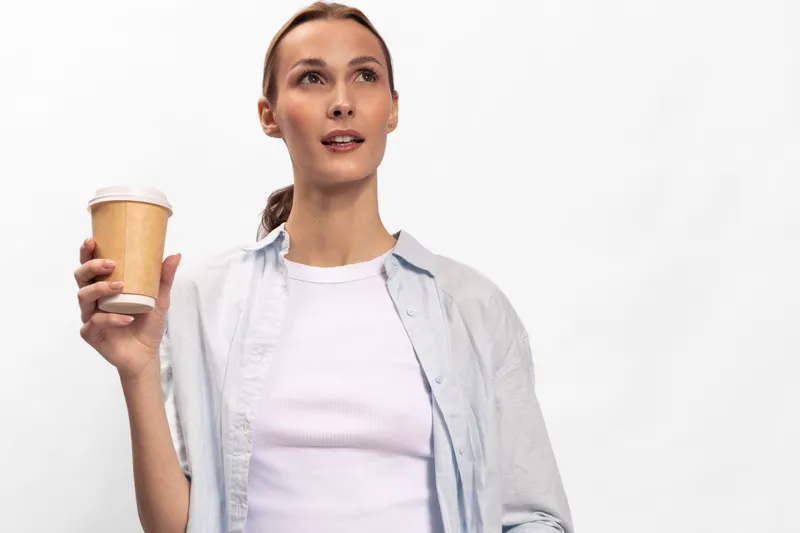 Young woman holding a disposable coffee cup looks away thoughtfully. Casual outfit and neutral background emphasize a relaxed moment of coffee break and daily routine concept.