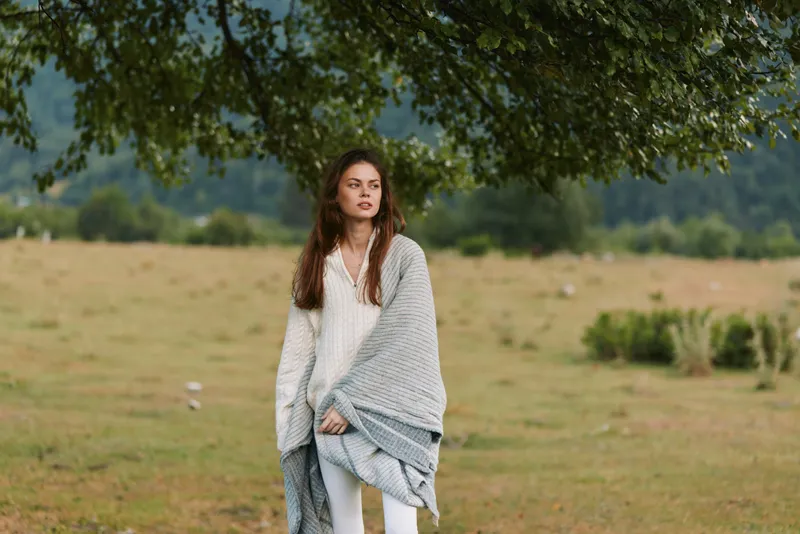 Woman in a gray shawl stands in an open field, natural lifestyle portrait, calm mood, soft light, outdoor countryside setting, serene and relaxed, she gazes into distance, casual pose, windswept