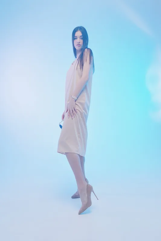 Young woman in a beige dress poses confidently in high heels against a soft blue background. Fashion and beauty concept with elegant style and modern minimalism.