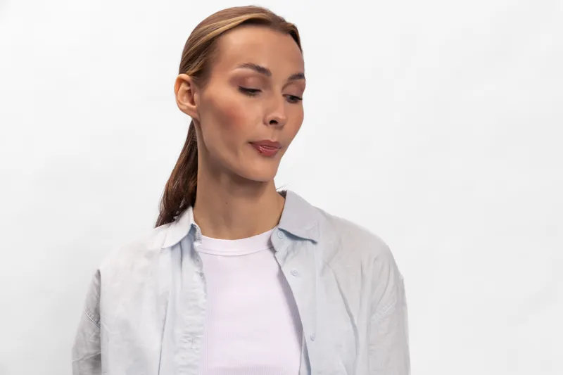 Young woman with long hair tied back wearing white shirt and light jacket looks down and to the side with closed lips on plain white background. Concept of contemplation and subtle emotion.