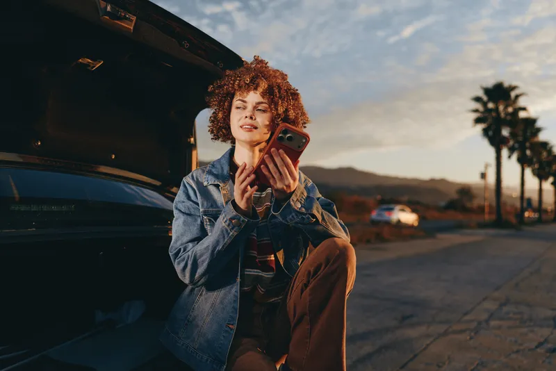 Woman lifestyle smartphone in hand near car on outdoor street at sunset, denim jacket, palm trees behind, relaxed communication mood, casual portrait, evening light, travel vibe