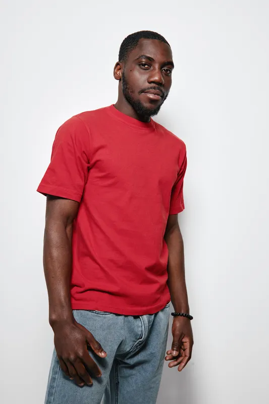 African American man with dark skin wearing red t shirt and blue jeans standing against white background looking at camera with confident expression and relaxed posture