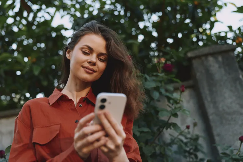 Bright outdoor woman lifestyle moment, smiling with phone in hand, casual red shirt, soft natural light, greenery background, candid and relaxed, everyday tech vibe soft