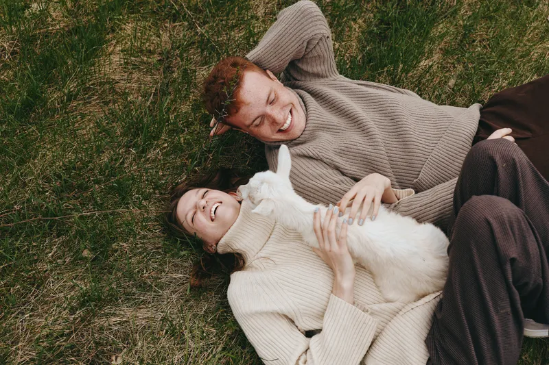 A relaxed couple lies on grass with a playful goat kid, enjoying outdoor life. The scene captures gentle affection, farm inspired calm, and a wholesome lifestyle moment in a sunny, natural setting.