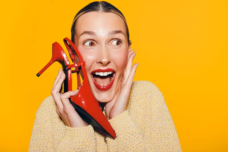 Excited woman with red lipstick holds red high heel shoes near face on bright yellow background. Concept of fashion, style, happiness, and vibrant emotions in studio lighting.
