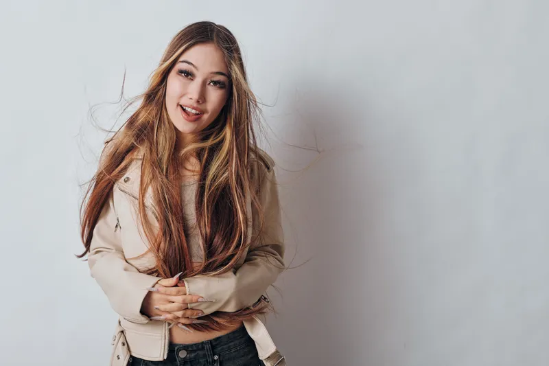 Young woman with long hair smiling, wearing beige jacket and jeans, standing against white background with hand gesture and natural expression, isolated studio portrait.