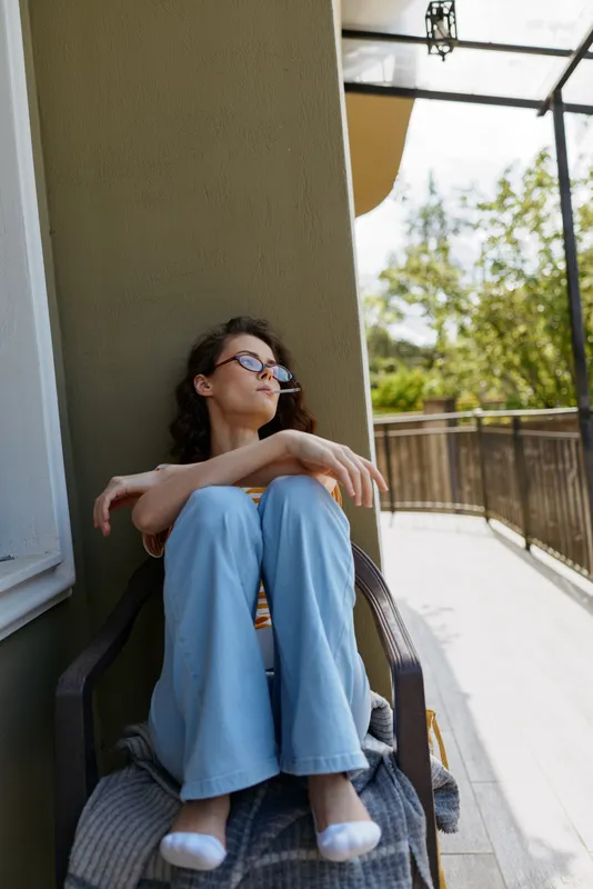 Relaxed woman sitting on a balcony in blue jeans and light top, sunlit outdoor space, calm mood, home life, casual comfort, peaceful self care, warm atmosphere and natural light warmth.