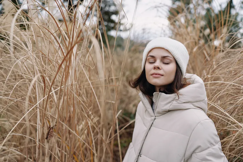 Woman in a light winter coat and white beanie stands among tall dry grasses in a calm outdoor winter scene with soft light and neutral tones.