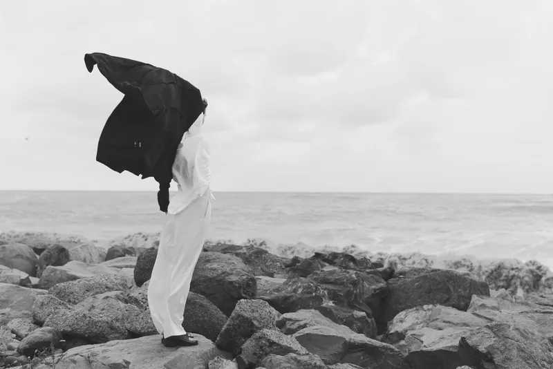 Silhouette, wind, jacket, beach, rocks, sea, motion, solitude, dramatic, outdoor scene capturing a figure lifting a jacket against gusts along a rocky shore in monochrome