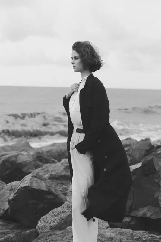 Coastal woman in a long coat stands on rocky shore by the ocean, wind swept hair and monochrome mood, a fashion scene capturing resilience and calm in a windy seascape