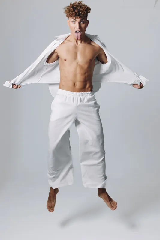 Young man jumping with shirt open showing bare chest and abs, playful expression with tongue out, wearing white pants on plain background in studio.